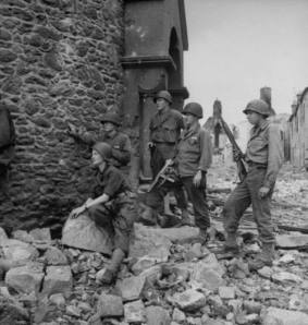 David E. Sherman: "Lee Miller con soldados americanos. Alemania, 1944" - La imagen muestra parte de una estructura en ruinas. Sobre los escombros, en primer término, aparece sentada Miller vestida con traje de campaña y casco. Detrás de ella, en pie, tres soldados norteamericanos armados vigilan mirando en la misma direcciín que ella. Pulse para ampliar.