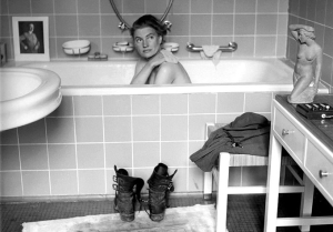David E. Sherman: "Lee Miller en la bañera de Hitler" (1945) - La imagen muestra un cuarto de baño limpio y grande. En primer término se aprecia a la derecha una consola son cajones sobre la que hay una escultura de una mujer desnuda agachada. A continuación hay un taburete sobre el que descansa un uniforme y, a su lado, una botas. A la izquierda aparece parte de un lavabo. Y al fondo, ocupando todo el ancho de baño, una gran bañera, con el frente alicatado, en la que se ve a Miller tomando un baño y pasándose una esponja por el hombro mientras mira a la cámara. Sobre la repisa del fondo aparecen varios soportes para poner jabón y una foto enmarcada de Adolf Hitler. Pulse para ampliar.