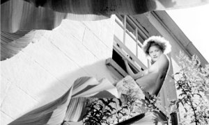 Roland Penrose - Fotografía de Lee Miller en Lamb Creek, Cornualles (1937) - La imagen muestra un plano contrapicado (tomado desde abajo) de Lee en lo alto de una escalera de madera en el lateral de una casa. Va desnuda de cintura para arriba y lleva una larga falda formada por una tela de flores que cae en pliegues alrededor de sus piernas. Pulse para ampliar.